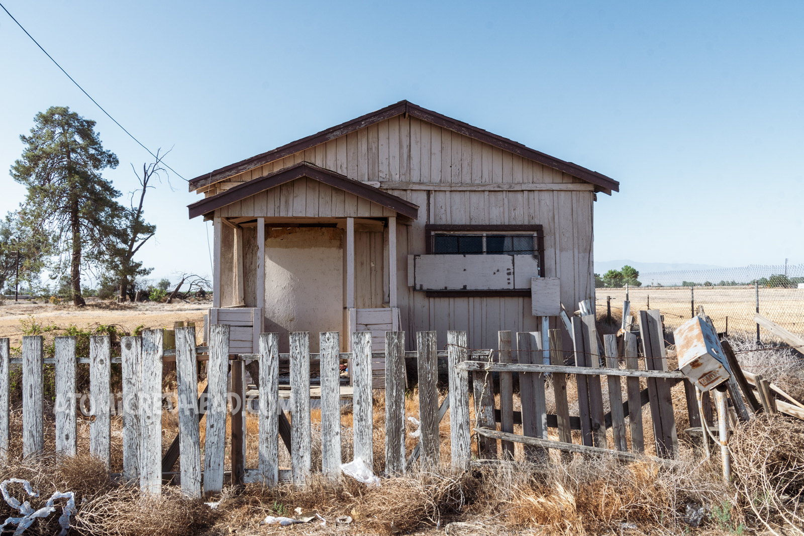 A small light brown home with dark brown trim, the front window is boarded up. A white picket fence with flaking paint sits in front.