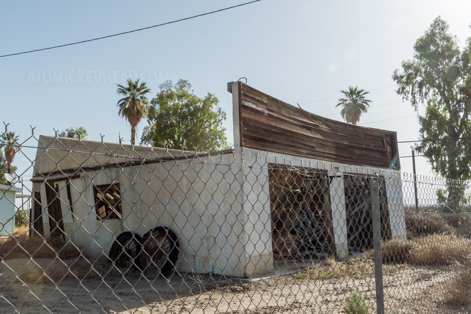 A stucco and wood garage style building, with the wooden roof that has collapsed in. Old wheels and tires are leaned against the side.