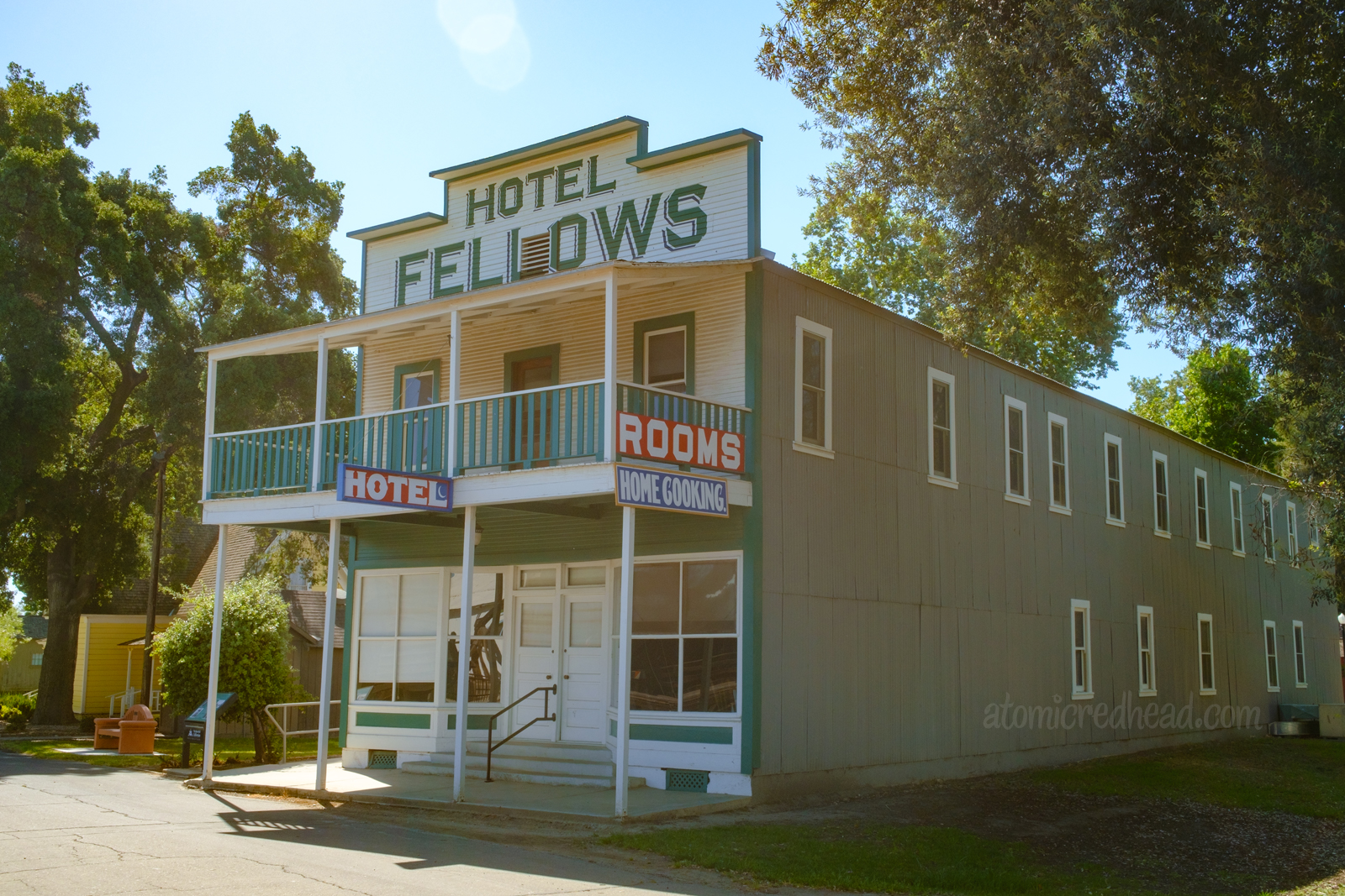 An old west style two story hotel, with balcony. Painted grey, with a white front, and blue trim. Along the top in large blue letters reads "Hotel Fellows" signs hanging from the balcony read "Rooms Home Cooking"
