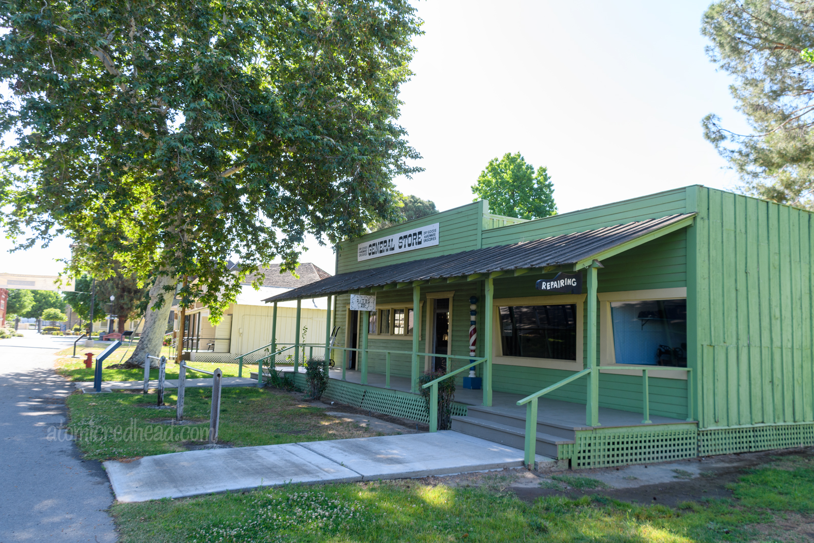 The General Store, painted green, with a porch. Two signs hang off the porch, one white with a bathtub painted on it reading "Baths 5 Cents" another sign shaped like a shoe reads "Repairing"