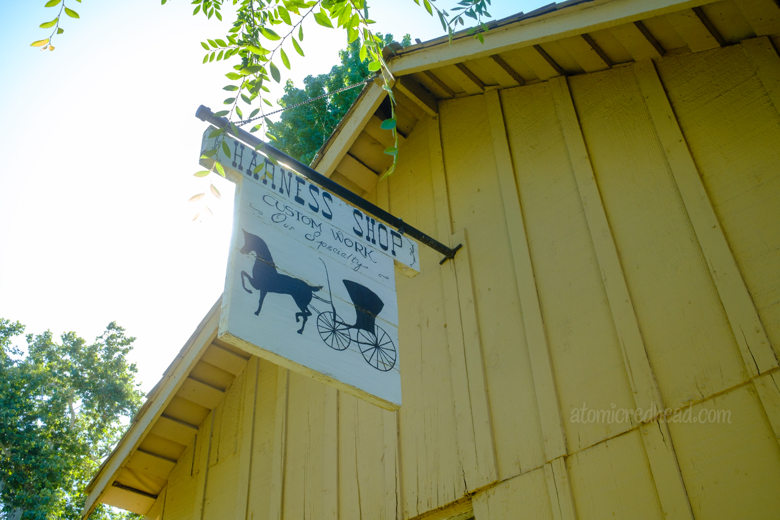 A white sign hangs from a yellow peaked roof, featuring a horse and buggy with script reading "Harness Shop Custom Work Our Specialty"