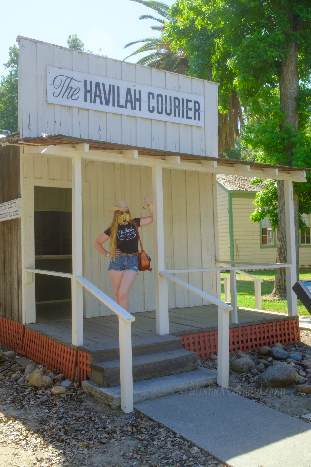 Myself standing on the porch of the newspaper building, an old west style building painted white with black letters at the top reading "The Havilah Courior", wearing a white cowboy hat, black shirt reading "Nudie's Rodeo Tailor", jean shorts, and a tooled leather purse that resembles a saddle.