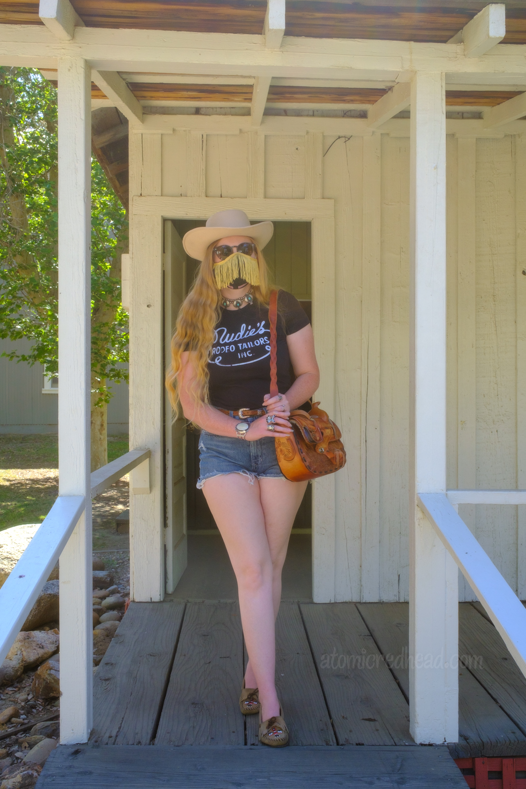 Myself standing on the porch of the newspaper building, wearing a white cowboy hat, black shirt reading "Nudie's Rodeo Tailor", jean shorts, and a tooled leather purse that resembles a saddle.