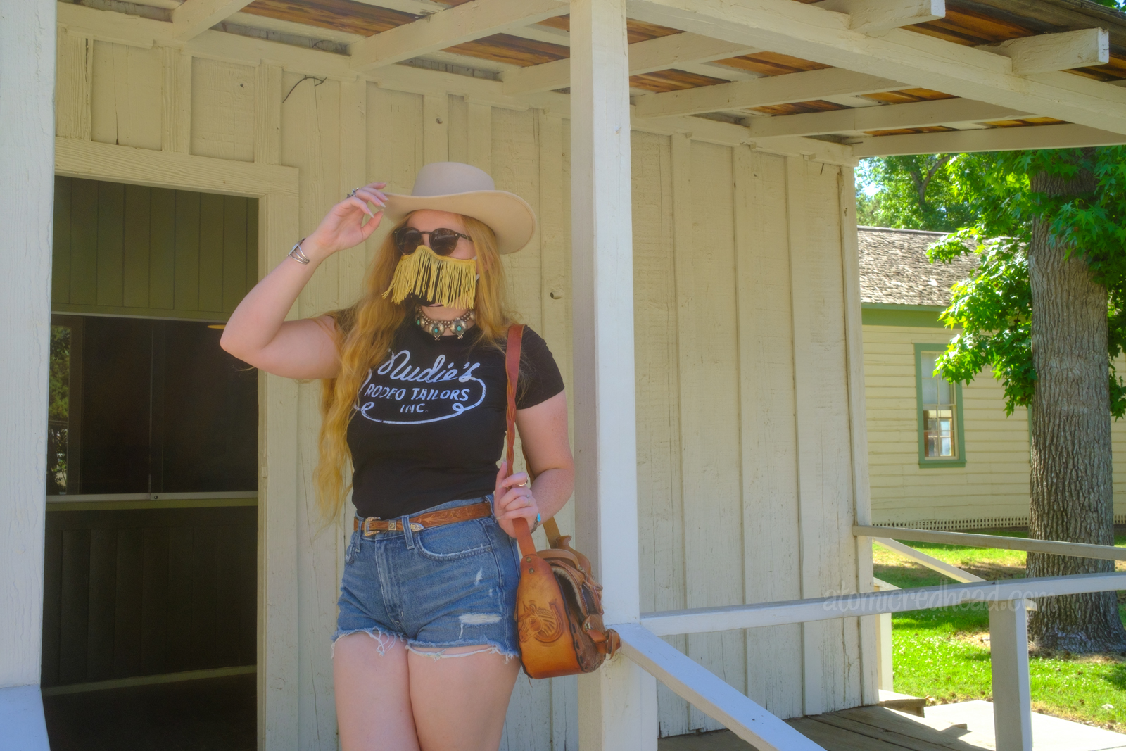 Myself standing on the porch of the newspaper building, wearing a white cowboy hat, black shirt reading "Nudie's Rodeo Tailor", jean shorts, and a tooled leather purse that resembles a saddle.