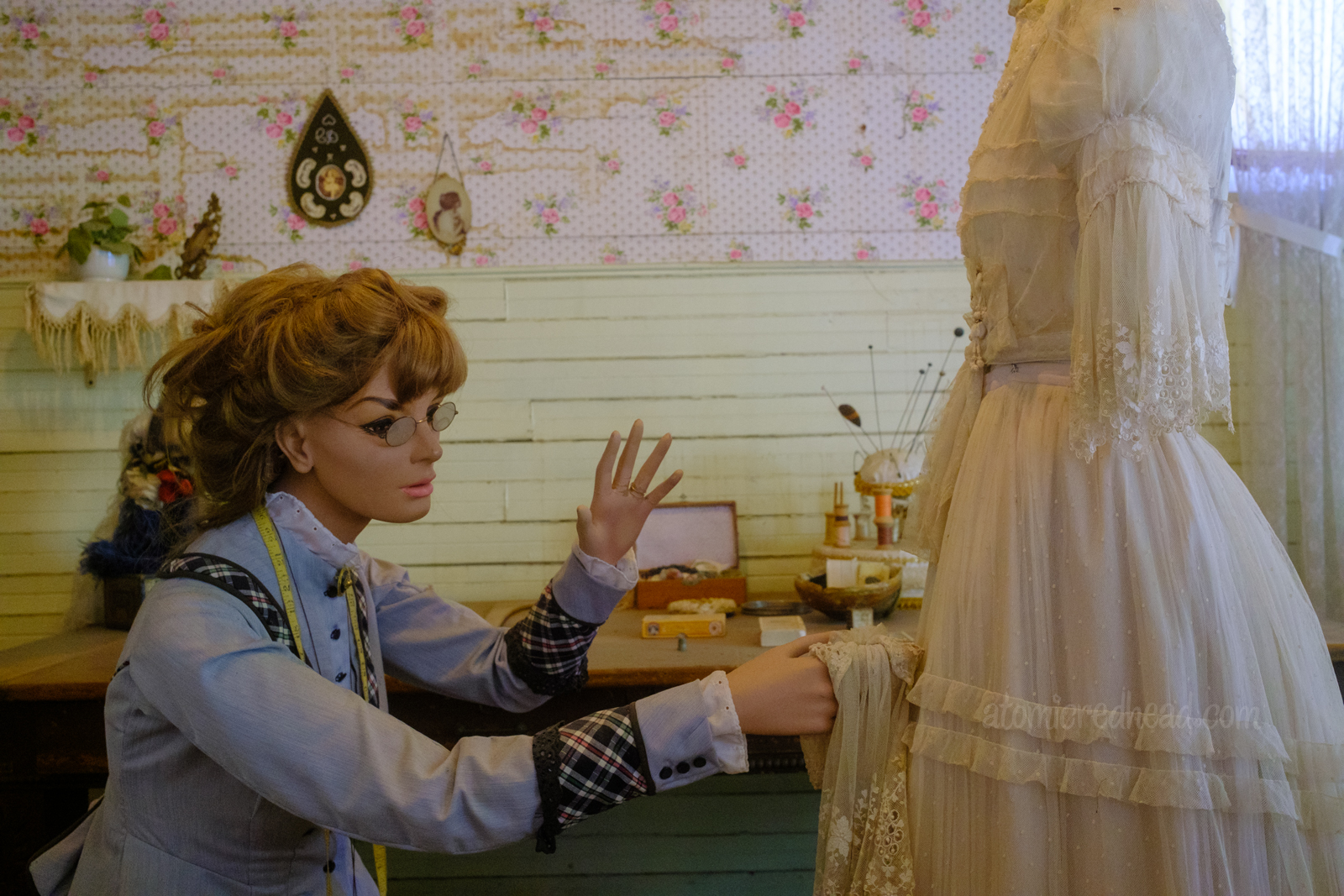 A mannequin of a woman appears as if she is working on a white dress that sits on a dressform.