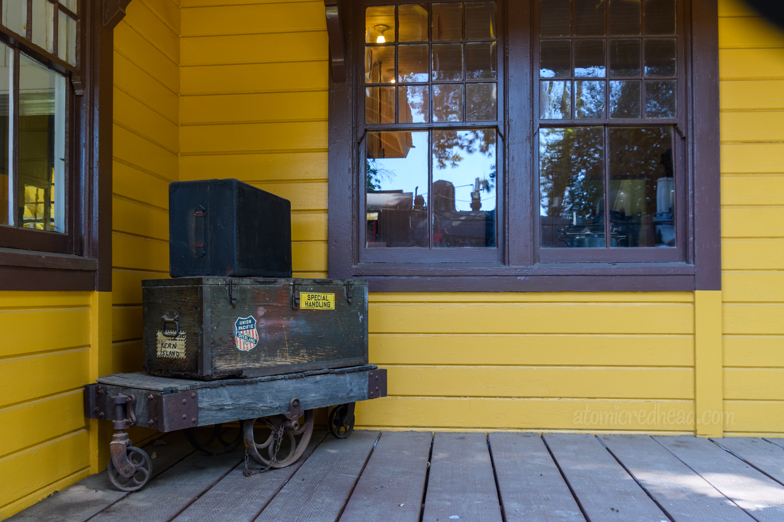 Old trucks sit on the platform of an old railroad station, which is painted a sunflower yellow with brown trim.