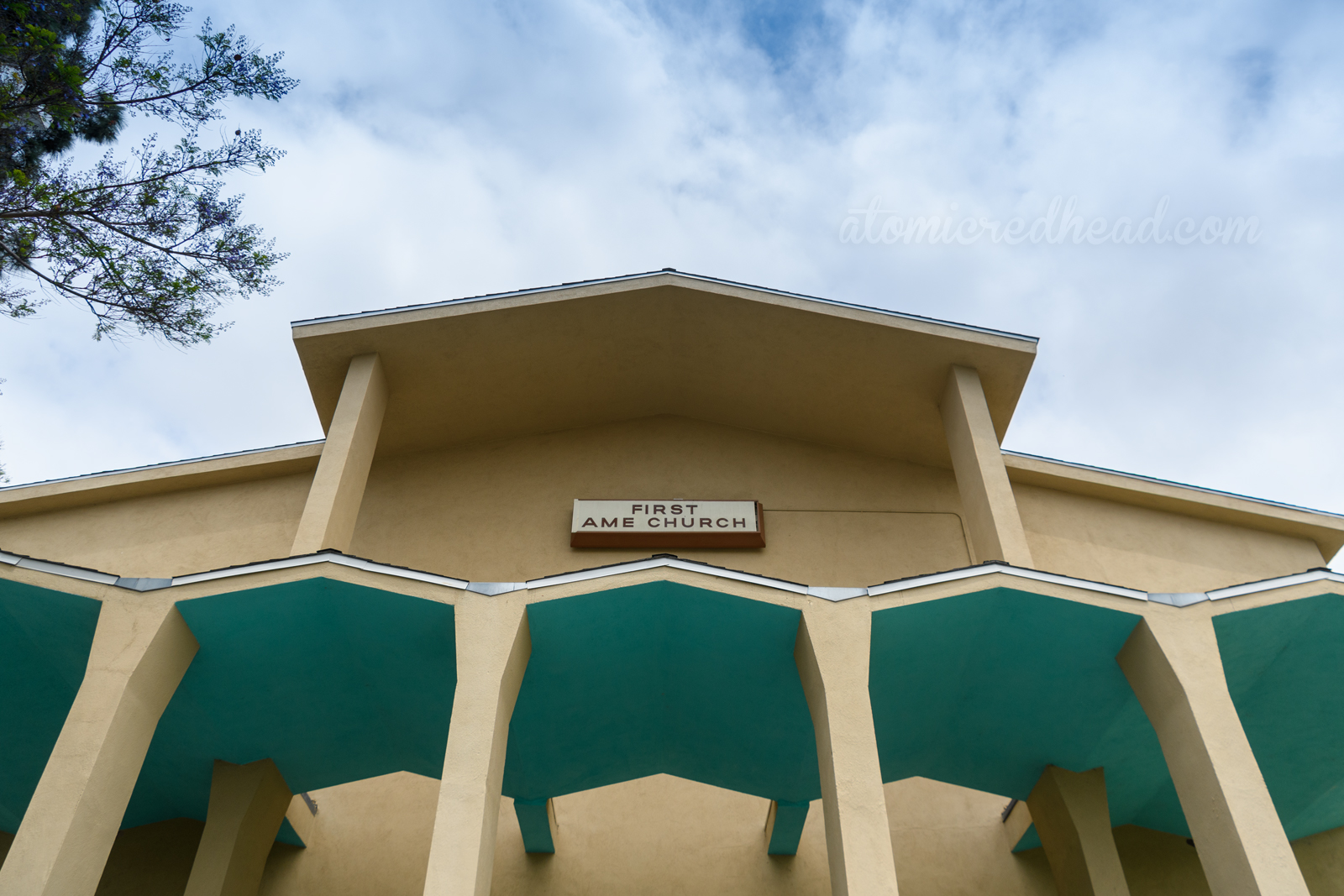 A pale yellow church with zig-zag roof that juts out over the doors, the underside painted turquoise. The sign reads "First AME Church"