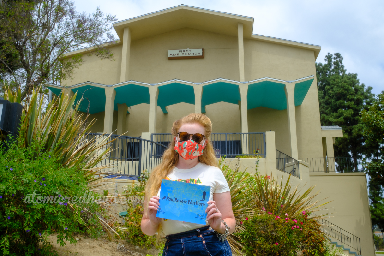 Myself in front of a pale yellow church with zig-zag roof that juts out over the doors, the underside painted turquoise. The sign reads "First AME Church" holding a sign that reads "#PaulRevereWasHere"