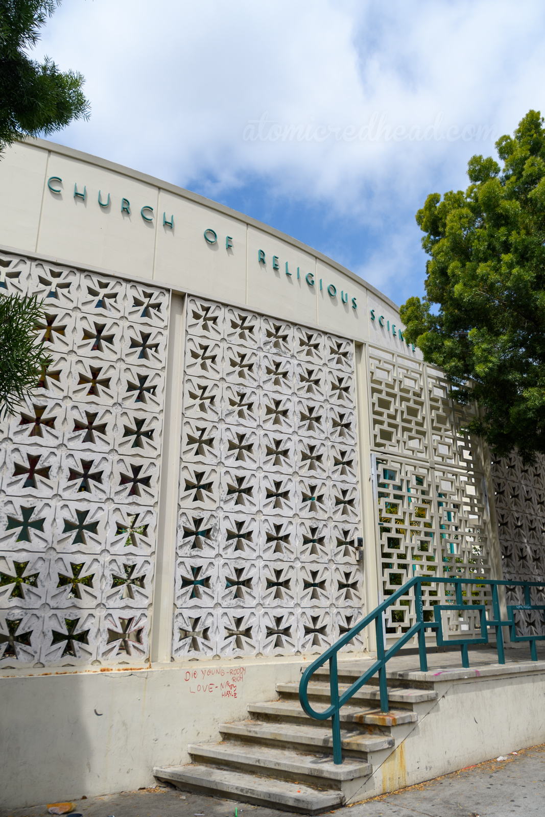 A white circular building sits behind breeze blocks with cut outs of a cross in the middle. Green letters along the top of the building read "Church of Religious Science"