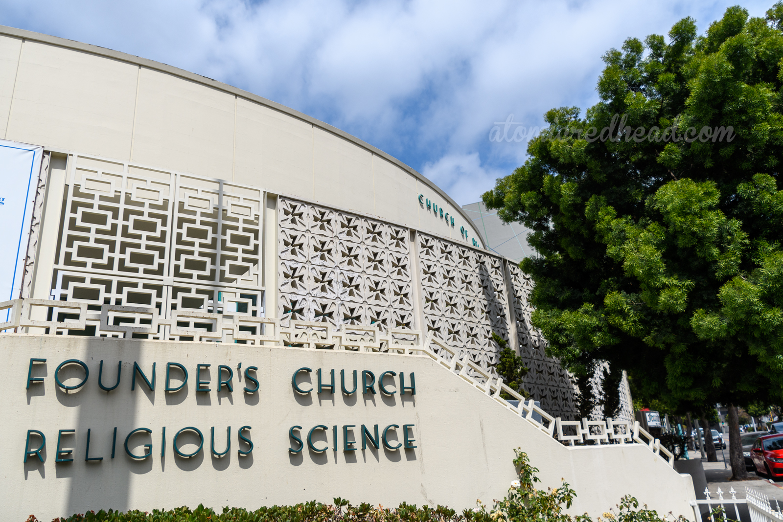 A white circular building sits behind breeze blocks with cut outs of a cross in the middle. Green letters are mounted along the staircase reading "Founders's Church Religious Science"