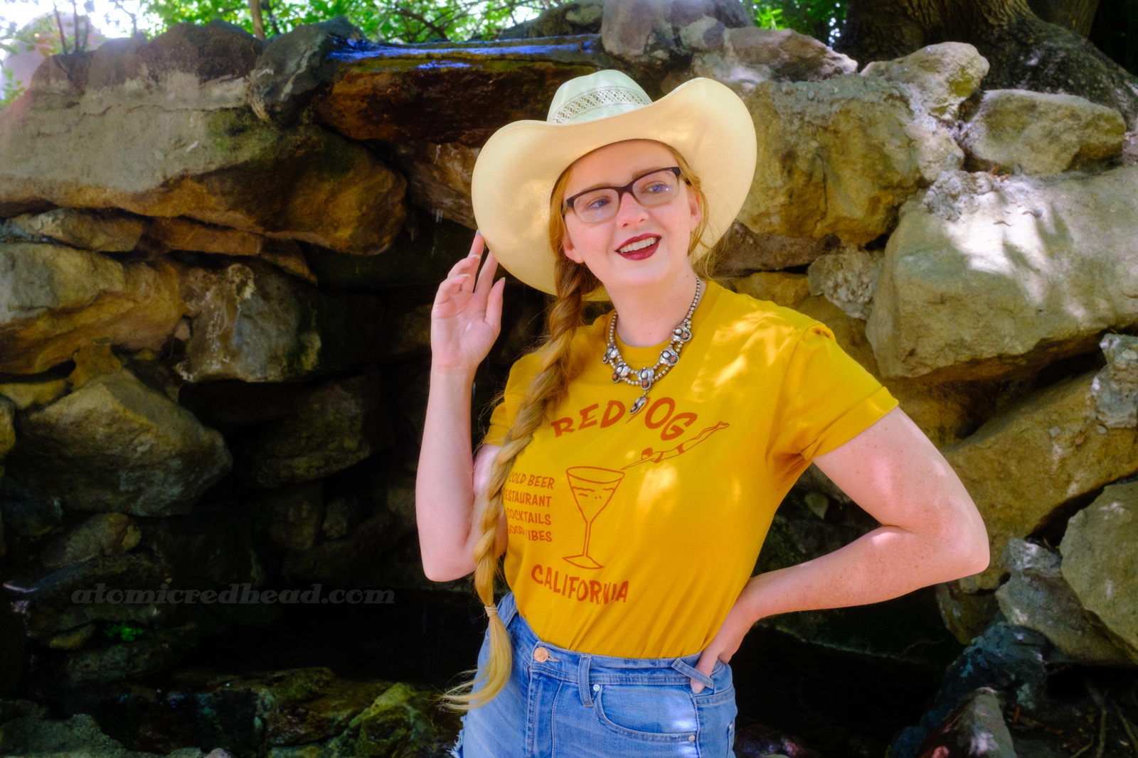 Myself, wearing a cream straw cowboy hat, yellow shirt red text reading "Red Dog California Cold Beer Restaurant Cocktails Good Vibes" and featuring a woman diving into a martini glass, and jean shorts, standing in front of a rock grotto.