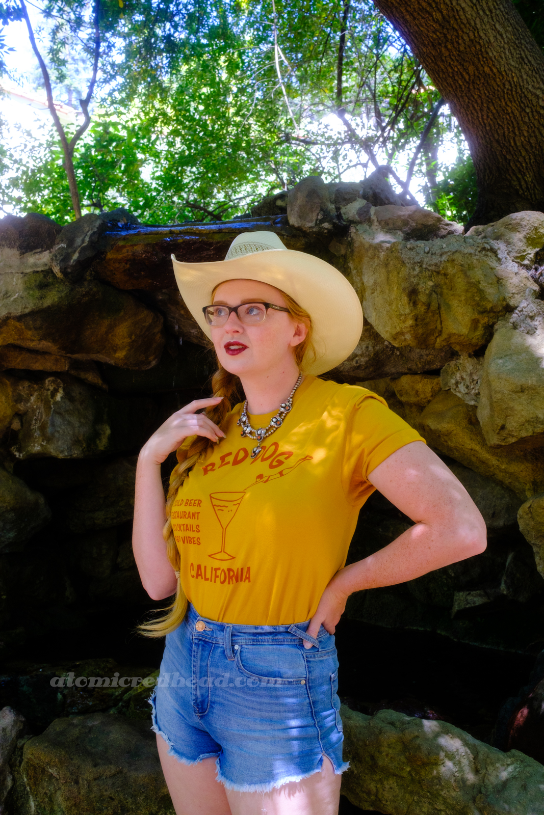 Myself, wearing a cream straw cowboy hat, yellow shirt red text reading "Red Dog California Cold Beer Restaurant Cocktails Good Vibes" and featuring a woman diving into a martini glass, and jean shorts, standing in front of a rock grotto.