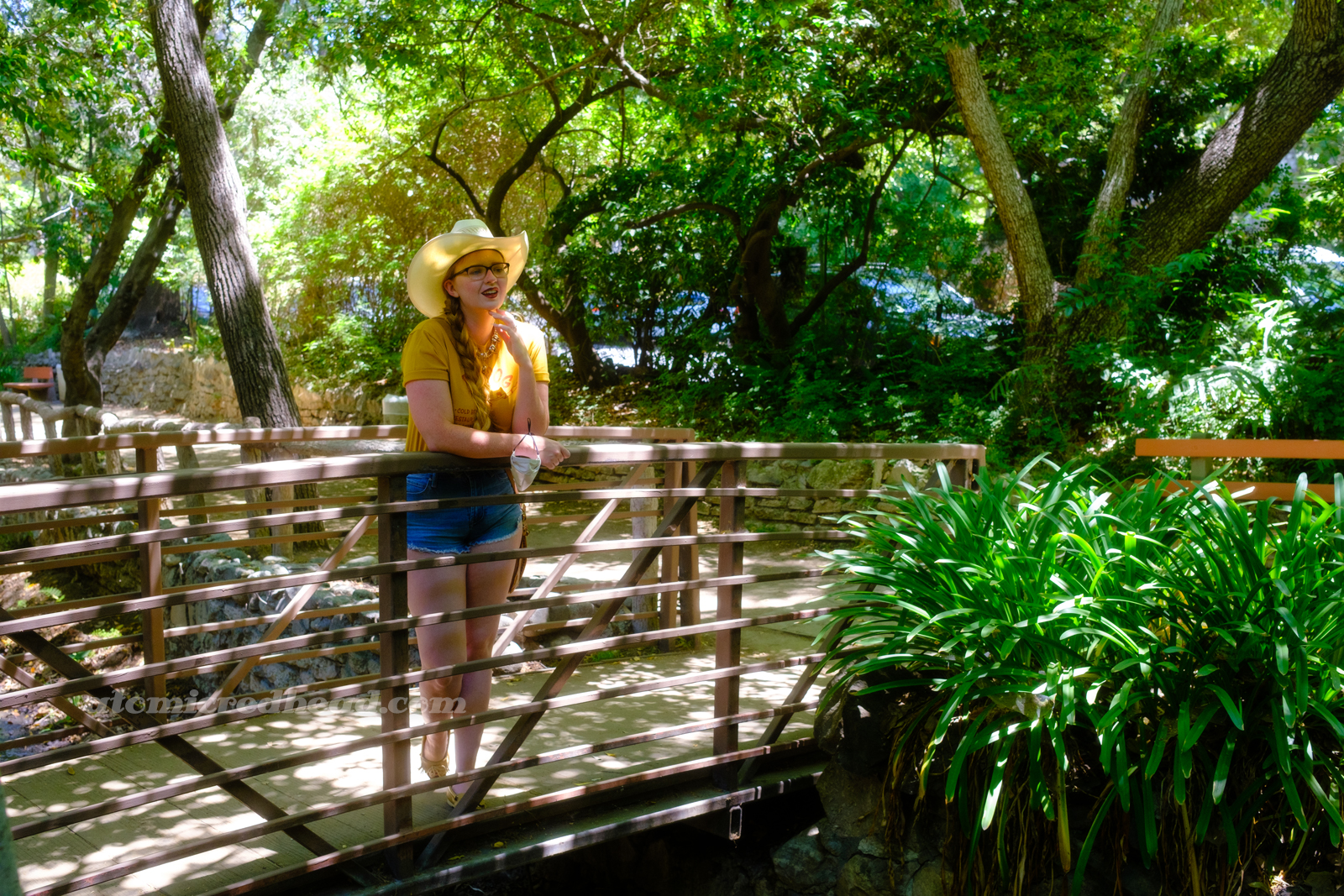 Myself, wearing a cream straw cowboy hat, yellow shirt red text reading "Red Dog California Cold Beer Restaurant Cocktails Good Vibes" and featuring a woman diving into a martini glass, and jean shorts, standing on a bridge over a small stream, surrounded by a variety of green plants.