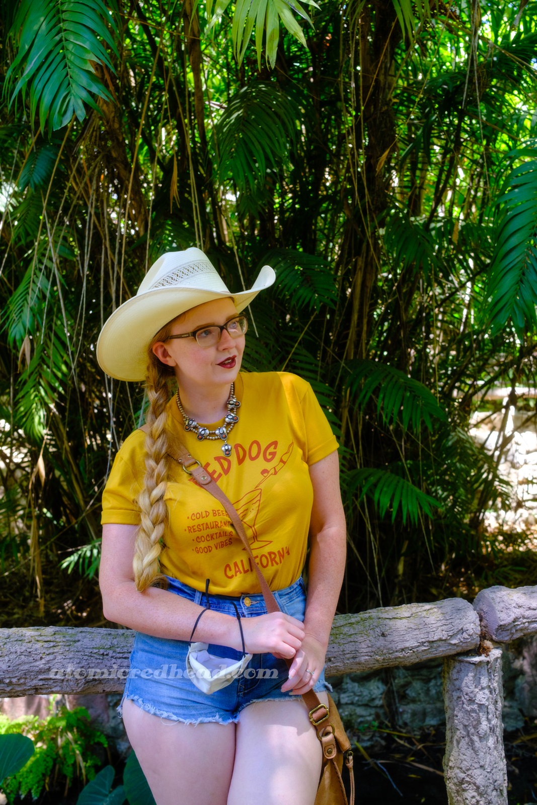 Myself, wearing a cream straw cowboy hat, yellow shirt red text reading "Red Dog California Cold Beer Restaurant Cocktails Good Vibes" and featuring a woman diving into a martini glass, and jean shorts, leaning against a railing made of concrete but made up to appear to be a log.