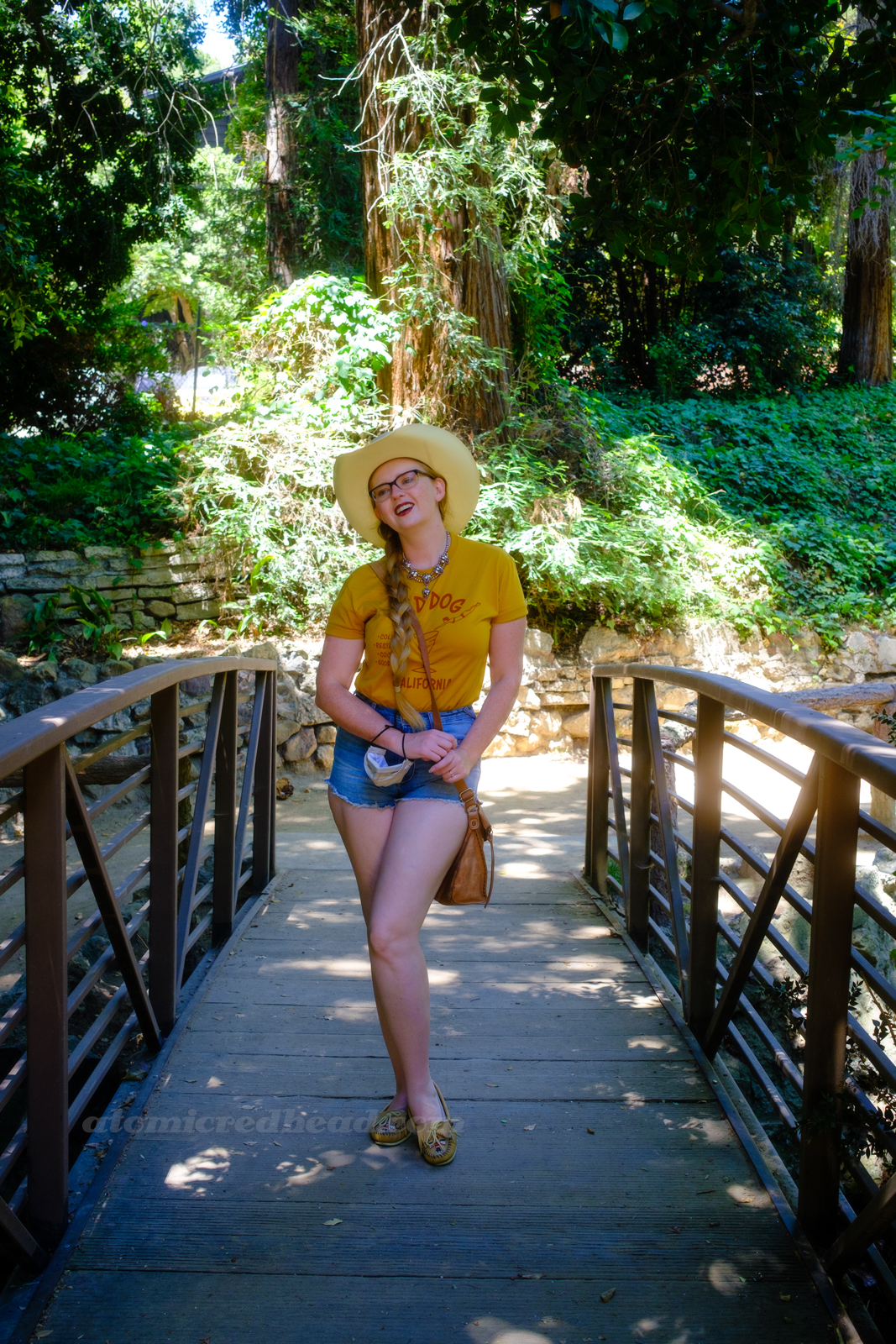 Myself, wearing a cream straw cowboy hat, yellow shirt red text reading "Red Dog California Cold Beer Restaurant Cocktails Good Vibes" and featuring a woman diving into a martini glass, and jean shorts, standing on a bridge over a small stream.