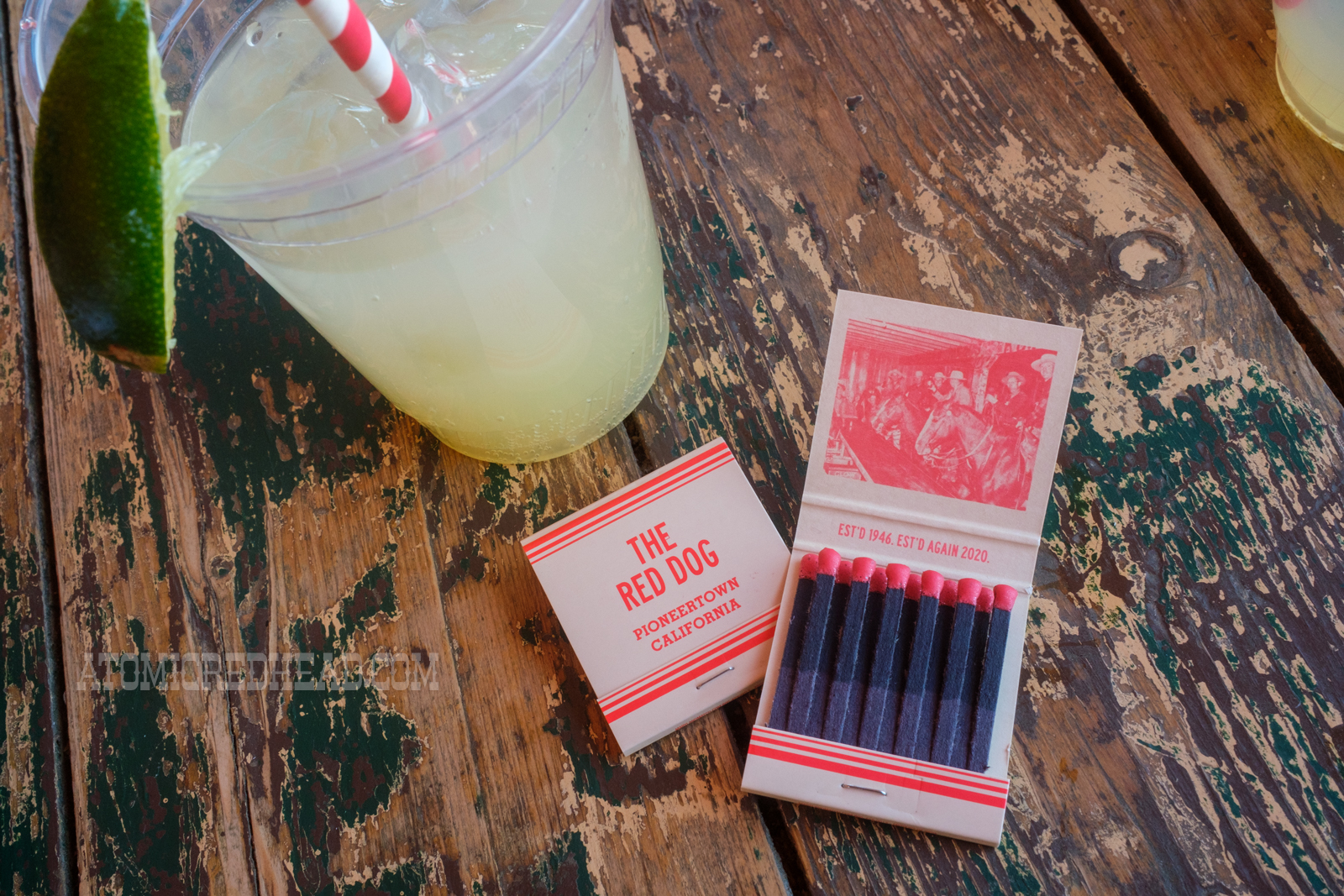 House made lemonade in a cup, and a pair of matchbooks. The closed matchbook reads "The Red Dog Pioneertown, California" the other is open and features an image of cowboys seated on horses at the bar. Text reads "Est'd 1946 Est'd Again 2020"