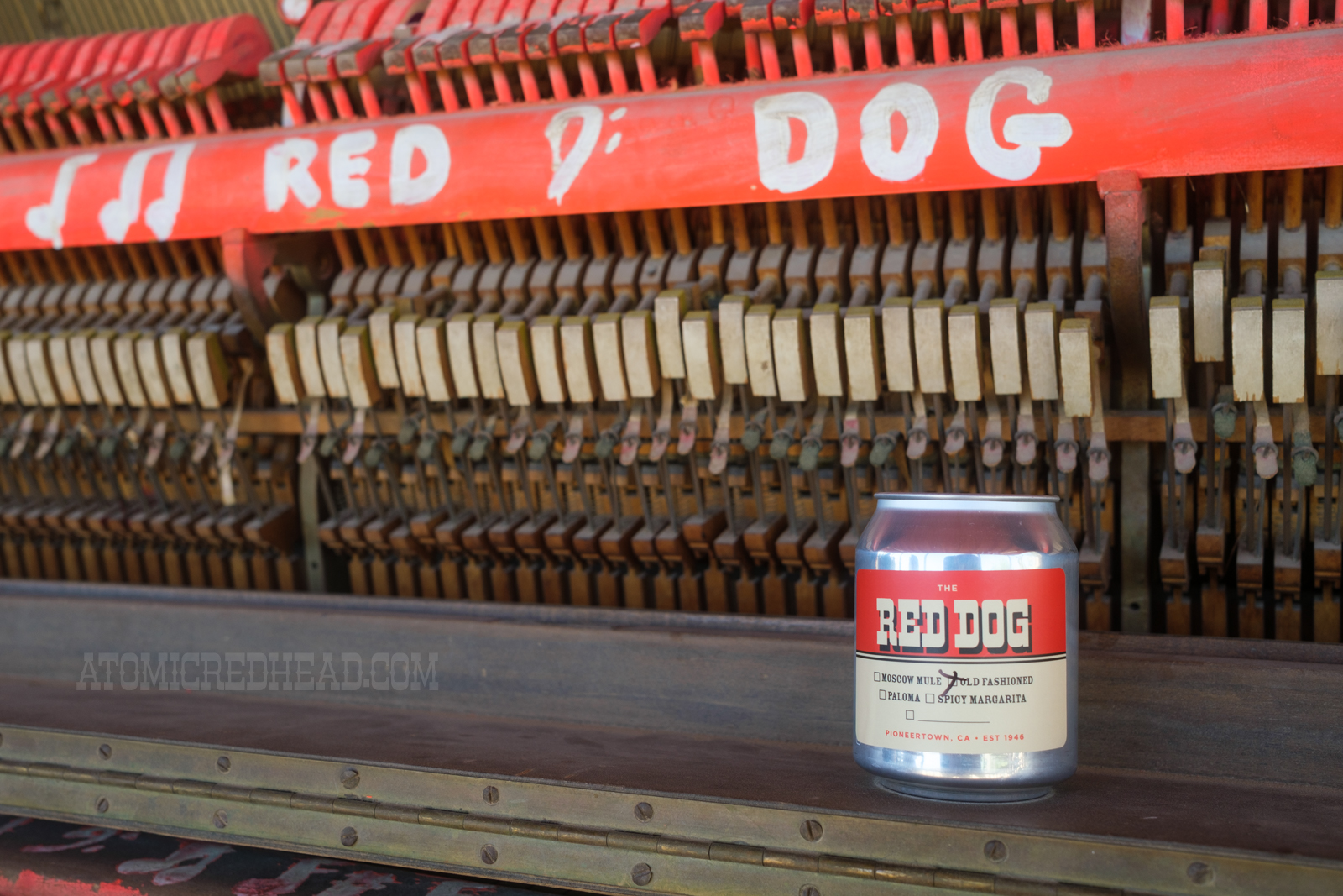 An old piano sits on the porch, painted on it reads "Red Dog" A small can reads "Red Dog" and a small check list below indicates which to-go cocktail is inside.