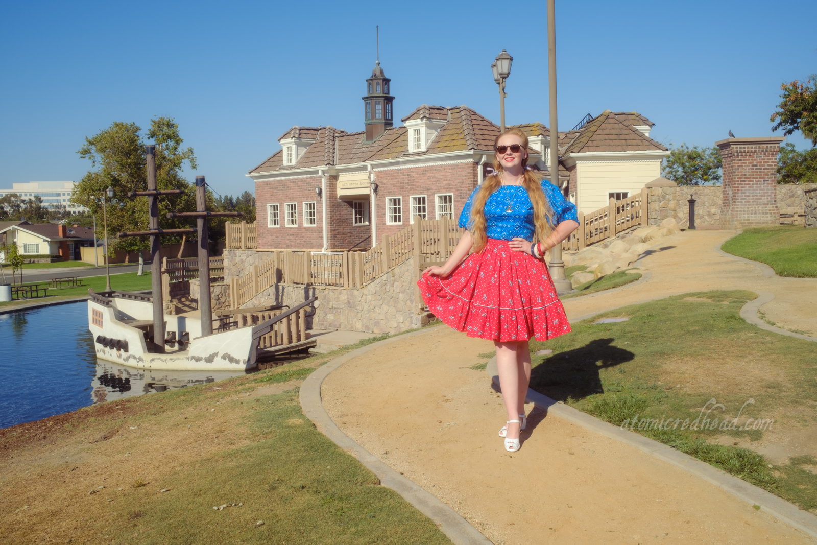 Myself, wearing a blue peasant top with a bandana print on it, a red skirt with "76" in white scattered throughout, and white shoes, standing in front of an old New England style building.
