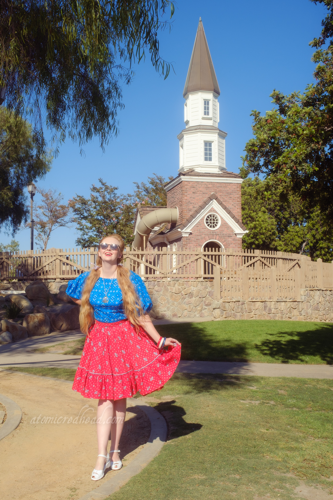 Myself, wearing a blue peasant top with a bandana print on it, a red skirt with "76" in white scattered throughout, and white shoes, standing in front of a small replica of the Old North Church.