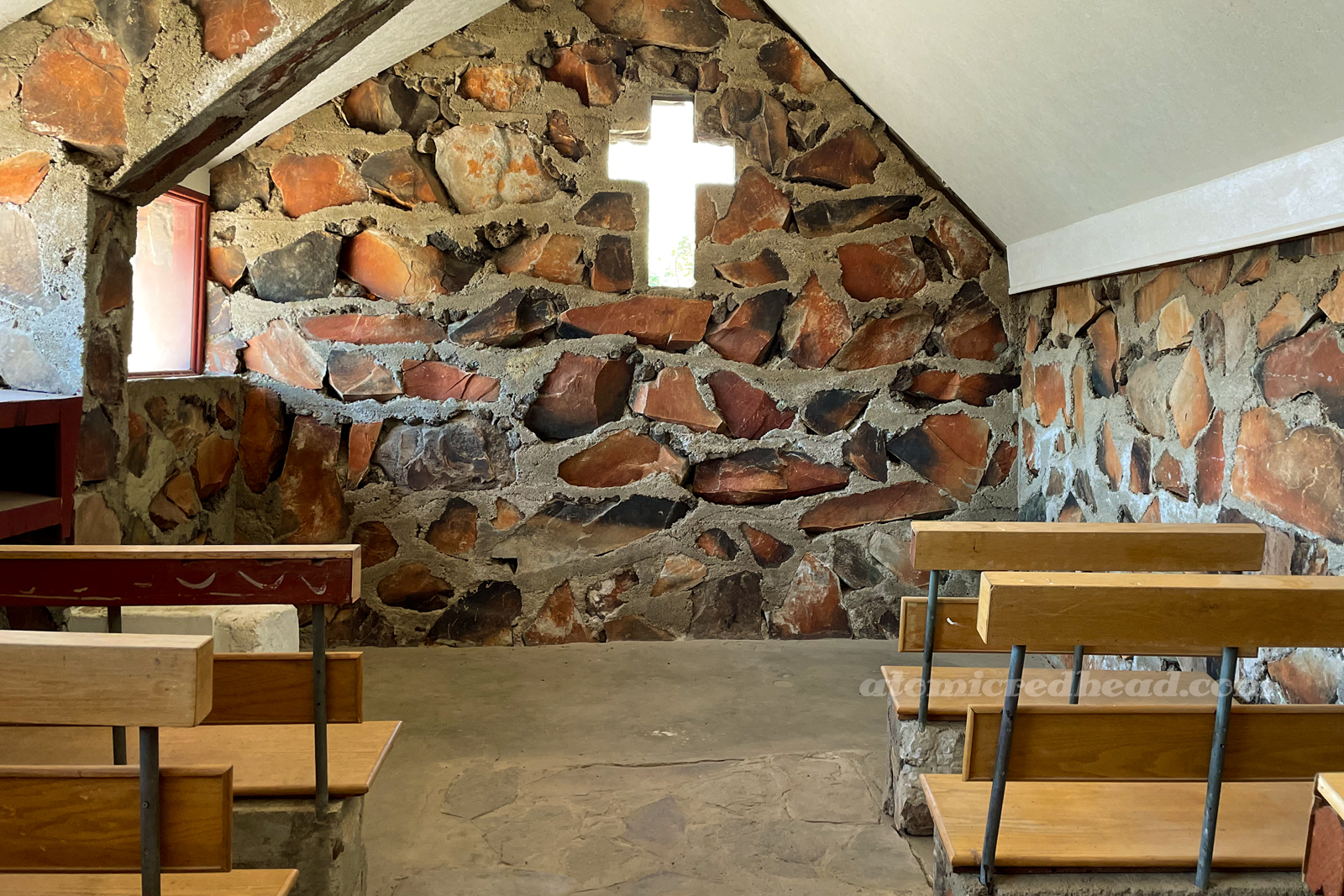 Inside the small stone church. The back wall features the warm, brown and orange rocks, with a small window shaped like a cross in the center. Wood benches flank the edges.