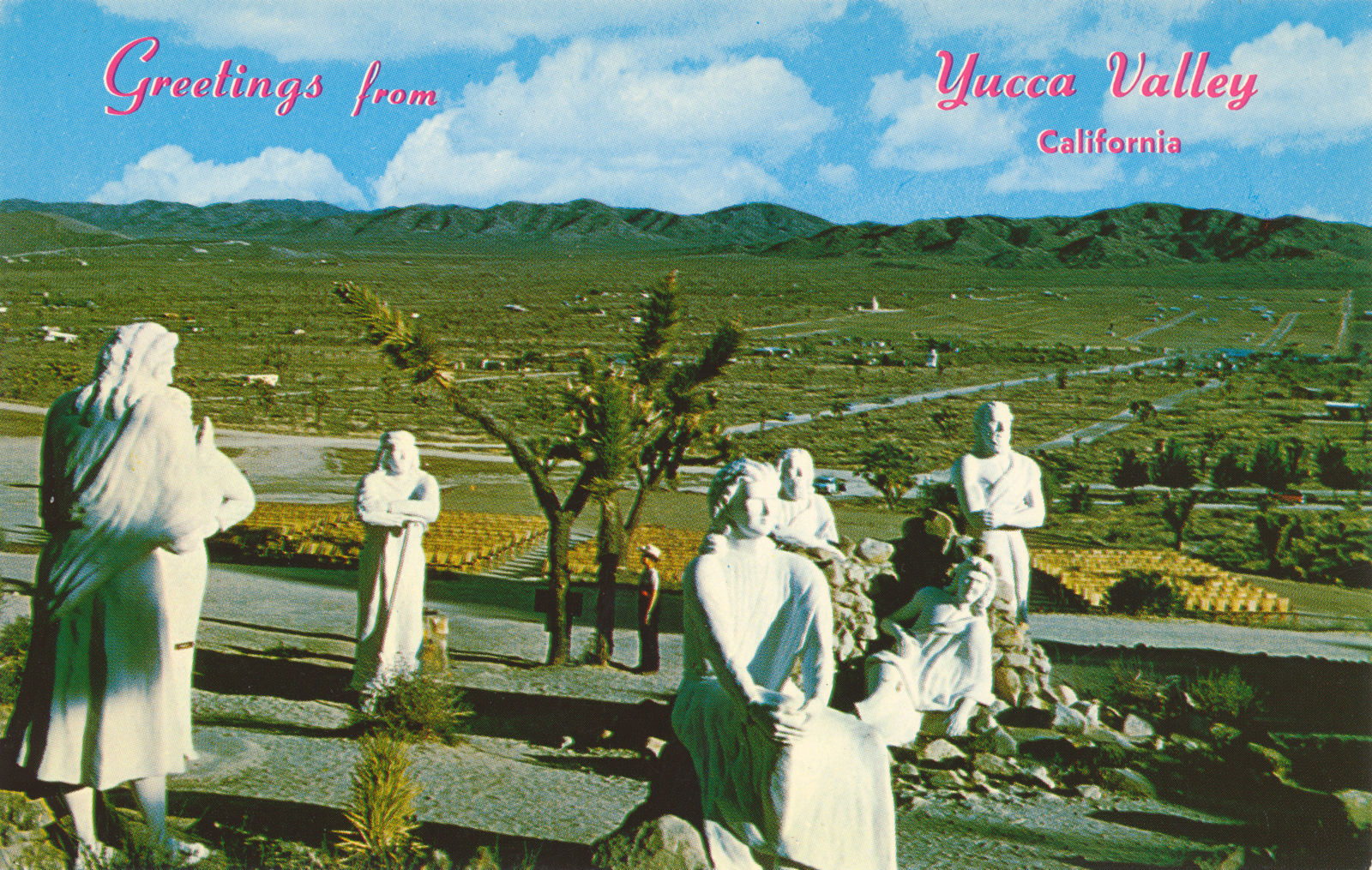 Vintage postcard that features an overview of Desert Christ Park with its white washed statues, Joshua trees, and the green expanse of a relatively undeveloped Yucca Valley in the distance. Red script reads "Greetings from Yucca Valley California"