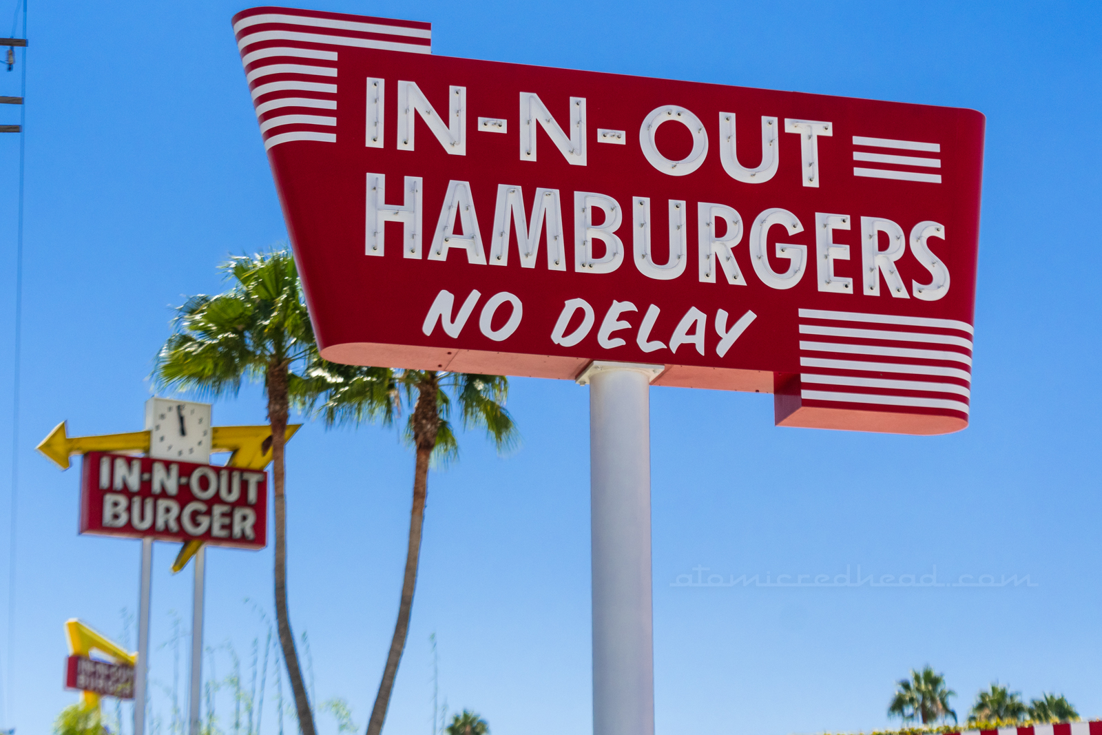 In the foreground is the replica of the original sign reading "In-N-Out Hamburgers No Delay" just beyond it is a later sign, featuring a yellow arrow and red rectangle reading "In N Out Burger" with a clock in the middle of the arrow, all of which features neon. And beyond that sign is the newest sign, red with a yellow arrow, reading "In N Out Burgers" but made of backlit plastic.
