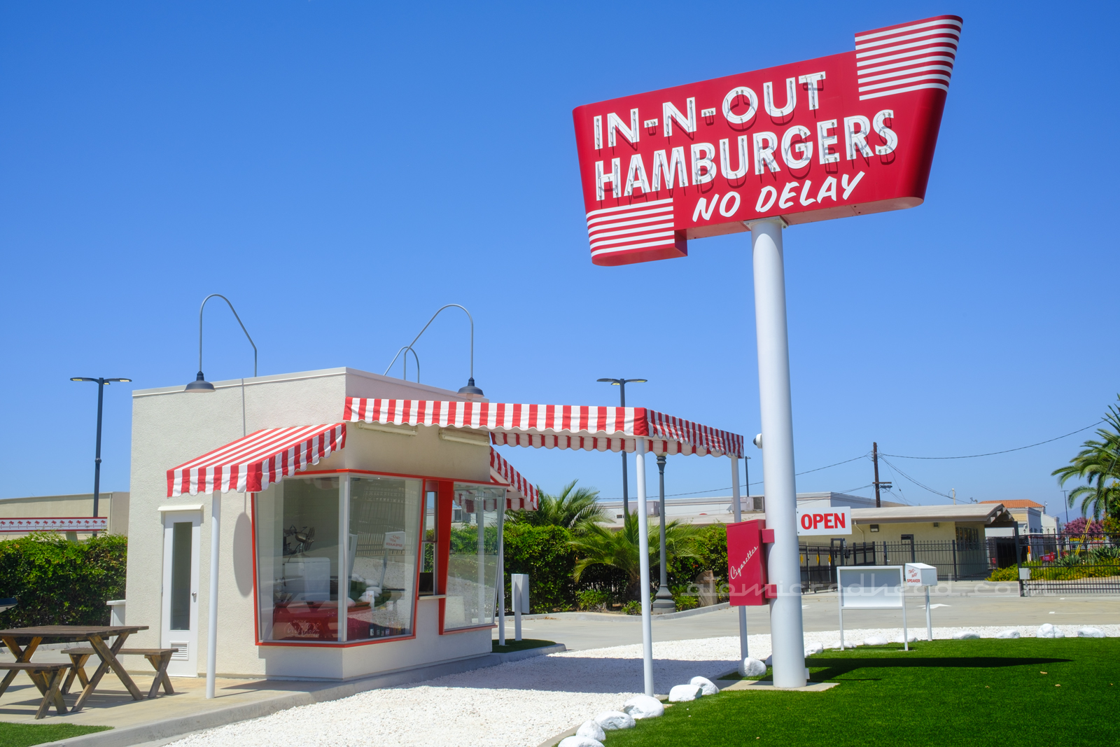 Replica of the first In-N-Out, a small, ten foot by ten foot white box, with red and white stripe awnings, a white rock horseshoe driveway curves up to the building. A tall sign to the right reads "In-N-Out Hamburger No Delay" in neon.
