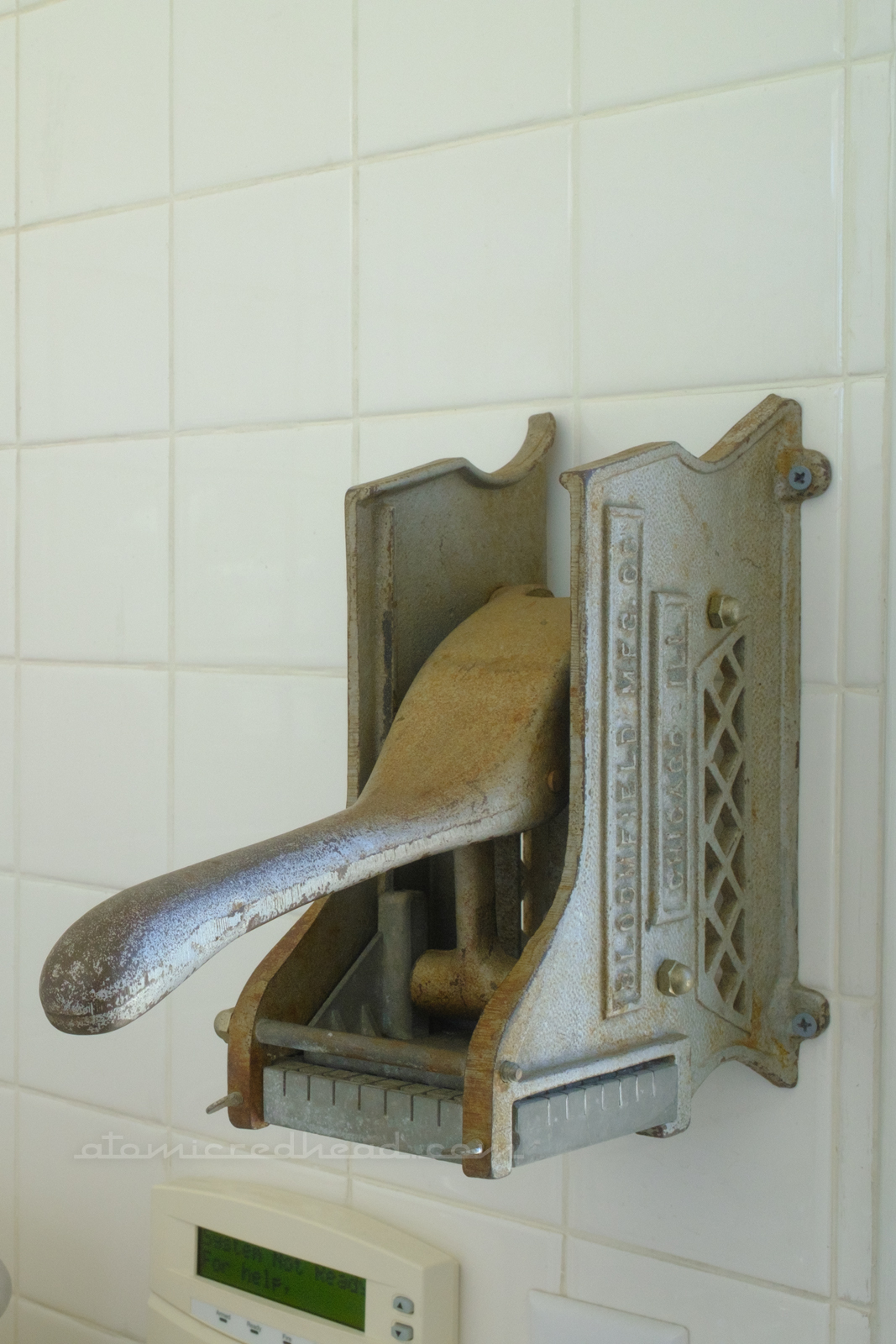 A vintage fry cutter mounted on a white tile wall.