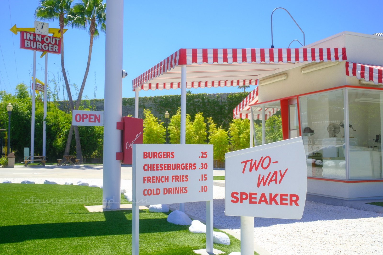 A white box reading "Two-Way Speaker" on it in red letters sits in the foreground, just beyond it is the menu board, reading "Burgers .25 Cheeseburgers .30 French Fries .15 Cold Drinks .10" in red letters, and just beyond it sits the replica building with a red and white stripe awning over the pick-up window.