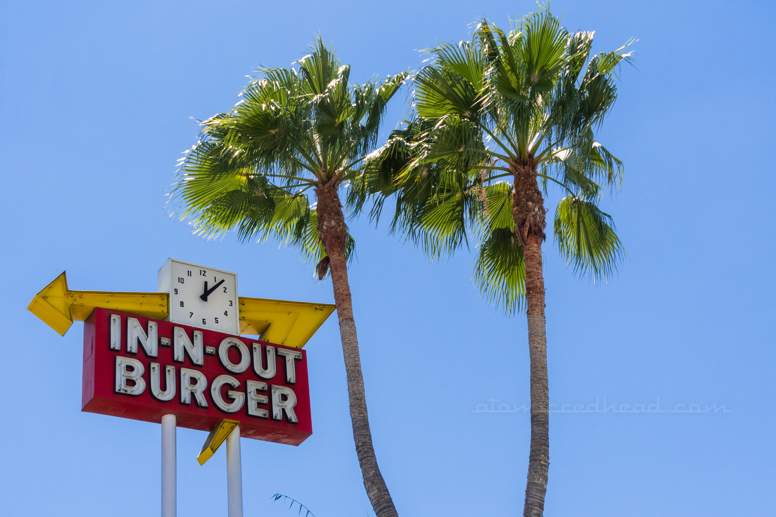 Close-up of a vintage neon In-N-Out sign with a clock incorporated into the sign. Crossed palm trees stand to the right.