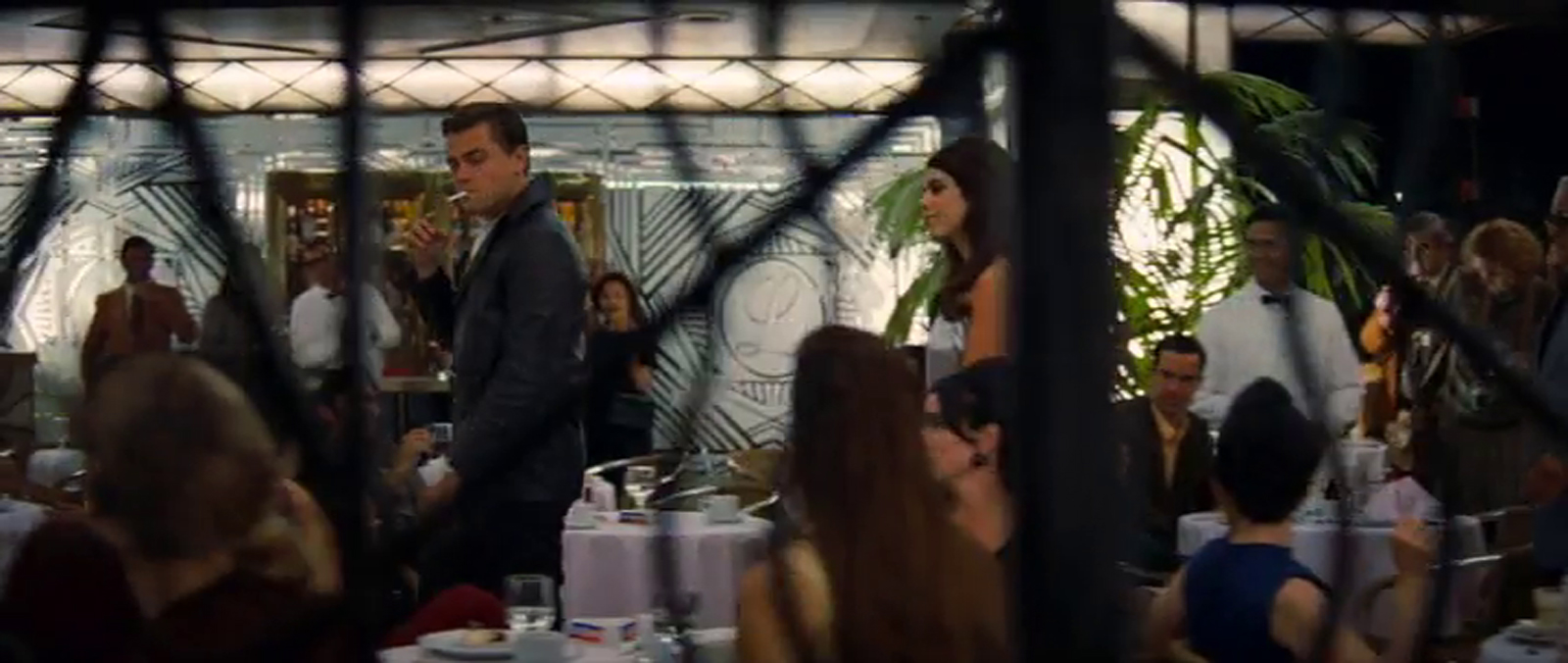 DiCaprio walks through the covered entryway of Cicada, which is posed as a restaurant, with various tables. The frosted glass and gold doors stand in the background.