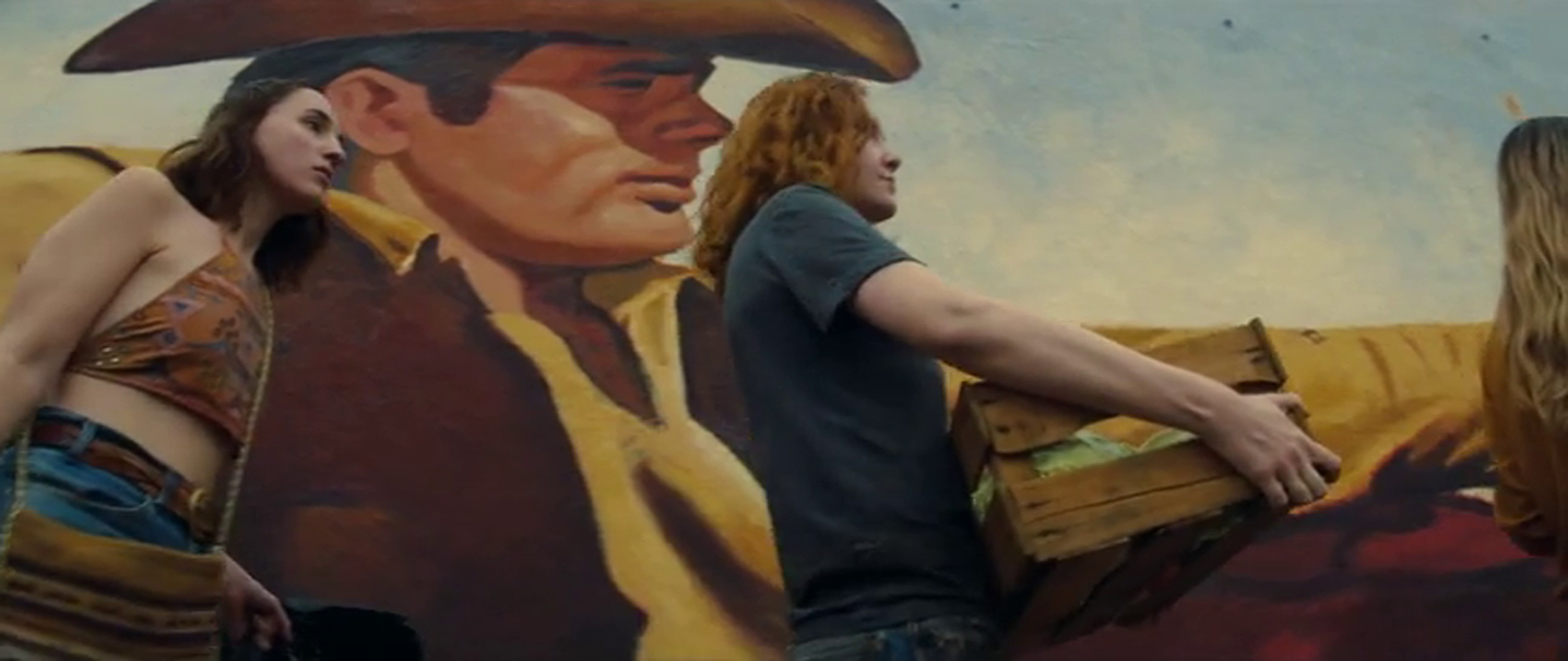 A massive mural of James Dean from his film Giant. He sits in a car dressed in cowboy attire, an old house visible in the distance, the hippie girls walk by carrying their dumpster finds.