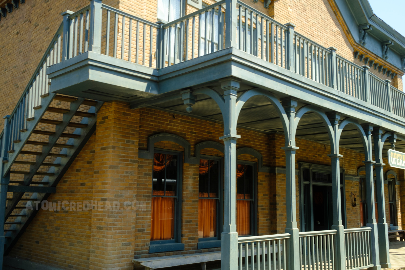 The saloon as it appears on Universal's backlot, a faux brick exterior with a dark green balcony and porch. A hanging sign out front reads "Opera House"