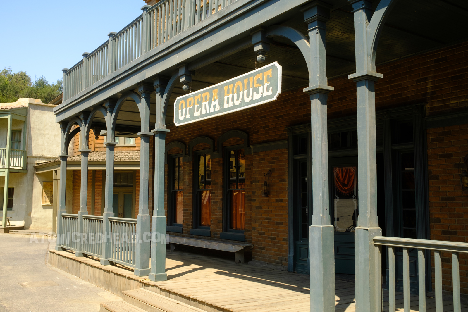 The saloon as it appears on Universal's backlot, a faux brick exterior with a dark green balcony and porch. A hanging sign out front reads "Opera House"