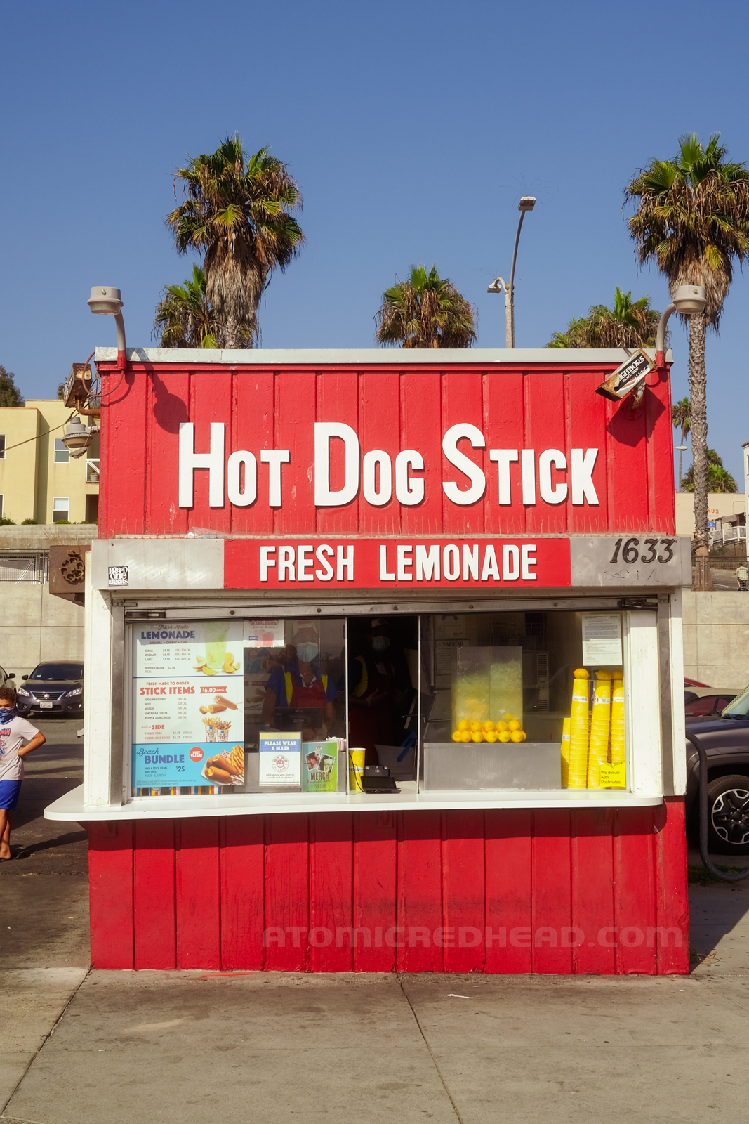 Front of the building, mainly painted red, with a white service window, and white letters reading "Hot Dog Stick Fresh Lemonade."