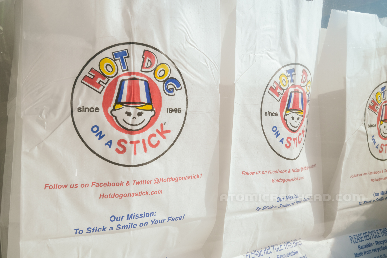 White paper bags await tasty goodies, the Hot Dog on a Stick logo printed in the middle, with text reading "Our Mission: To Stick a Smile on Your Face."