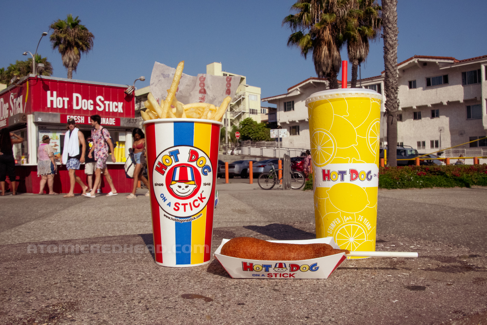 An early evening meal, French fries in a cup, a corndog and a tall cup of lemonade.