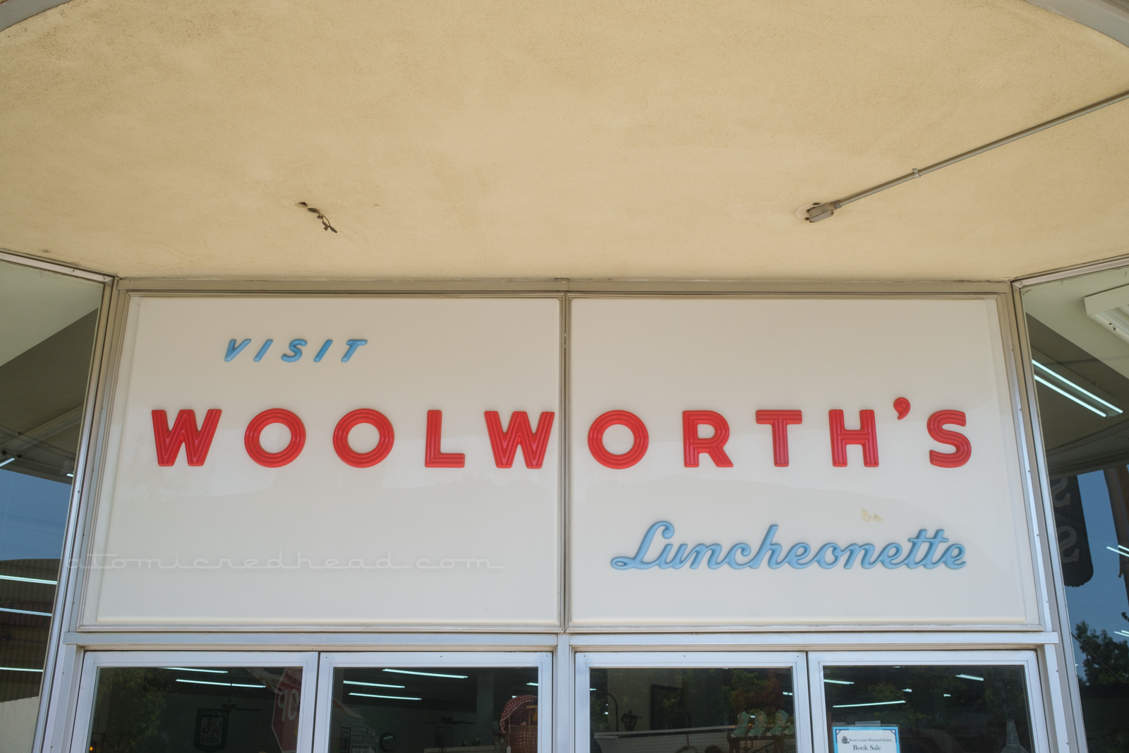 A large white plastic sign sits above the doorway to the antique mall. Small blue letters read "Visit" followed by large red letters reading "Woolorth's" and then below in smaller blue script reads "Luncheonette"