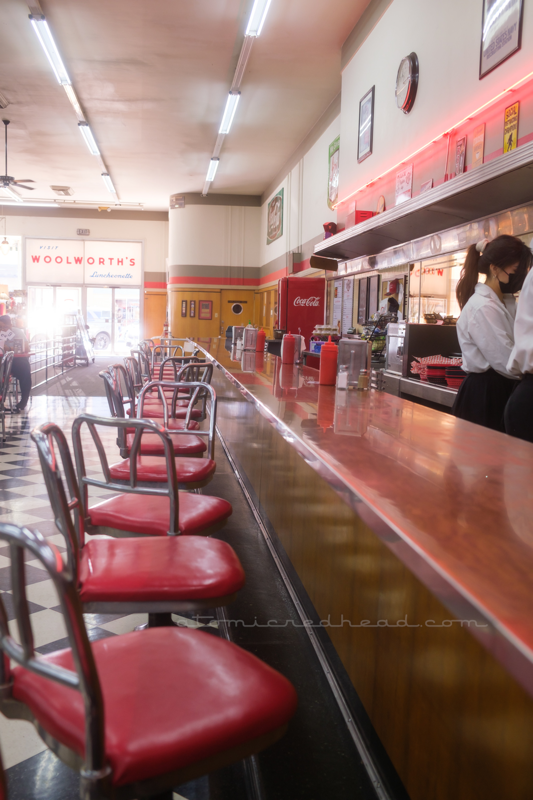 Looking down the lunch counter toward the front door. Pale wood paneled walls meet the tile backsplash of the work area behind the counter. Chrome backed stools also feature red upholstery and sit on top of black and white checkered floor.