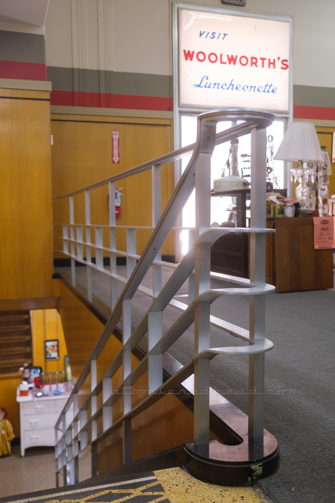 Swooping streamline hand rail curves toward the basement. A large white plastic sign read "Visit Woolworth's Luncheonette" in the background.