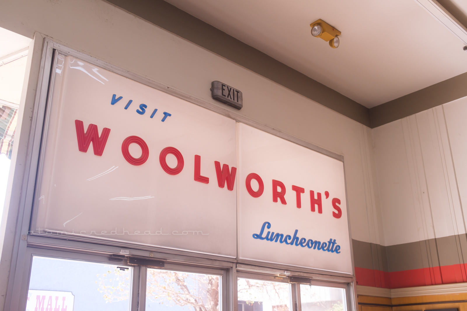 A large white plastic sign sits above the doorway to the antique mall. Small blue letters read "Visit" followed by large red letters reading "Woolorth's" and then below in smaller blue script reads "Luncheonette"