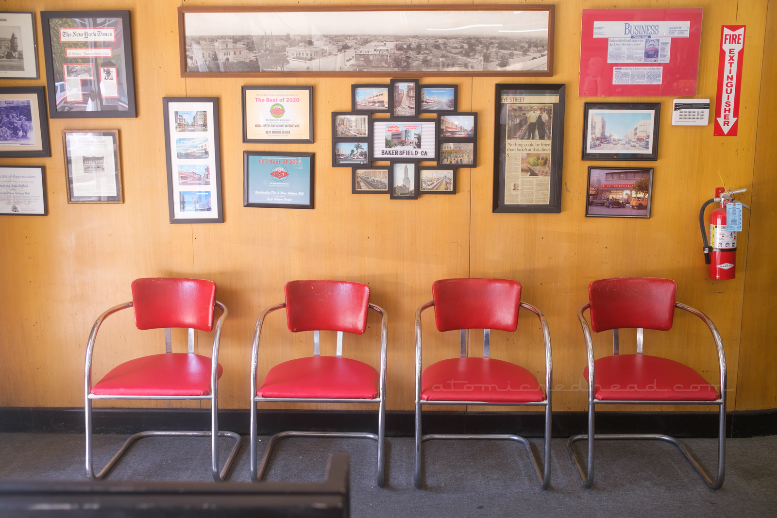 A pale wood paneled wall features framed articles, awards, and photos of the Woolworth's through the years, red upholstered and chrome chairs sit against the wall.