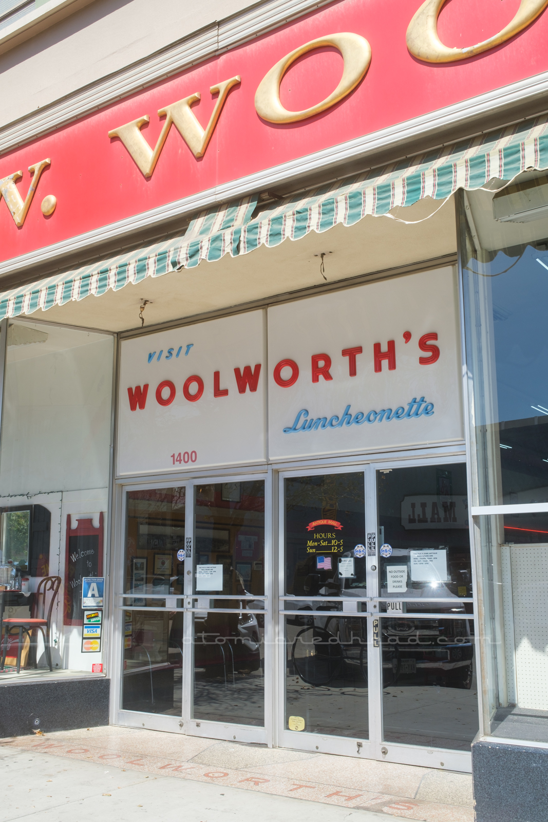 Under a red and gold sign read "F.W. Woolworth's" is a large white plastic sign sits above the doorway to the antique mall. Small blue letters read "Visit" followed by large red letters reading "Woolorth's" and then below in smaller blue script reads "Luncheonette"