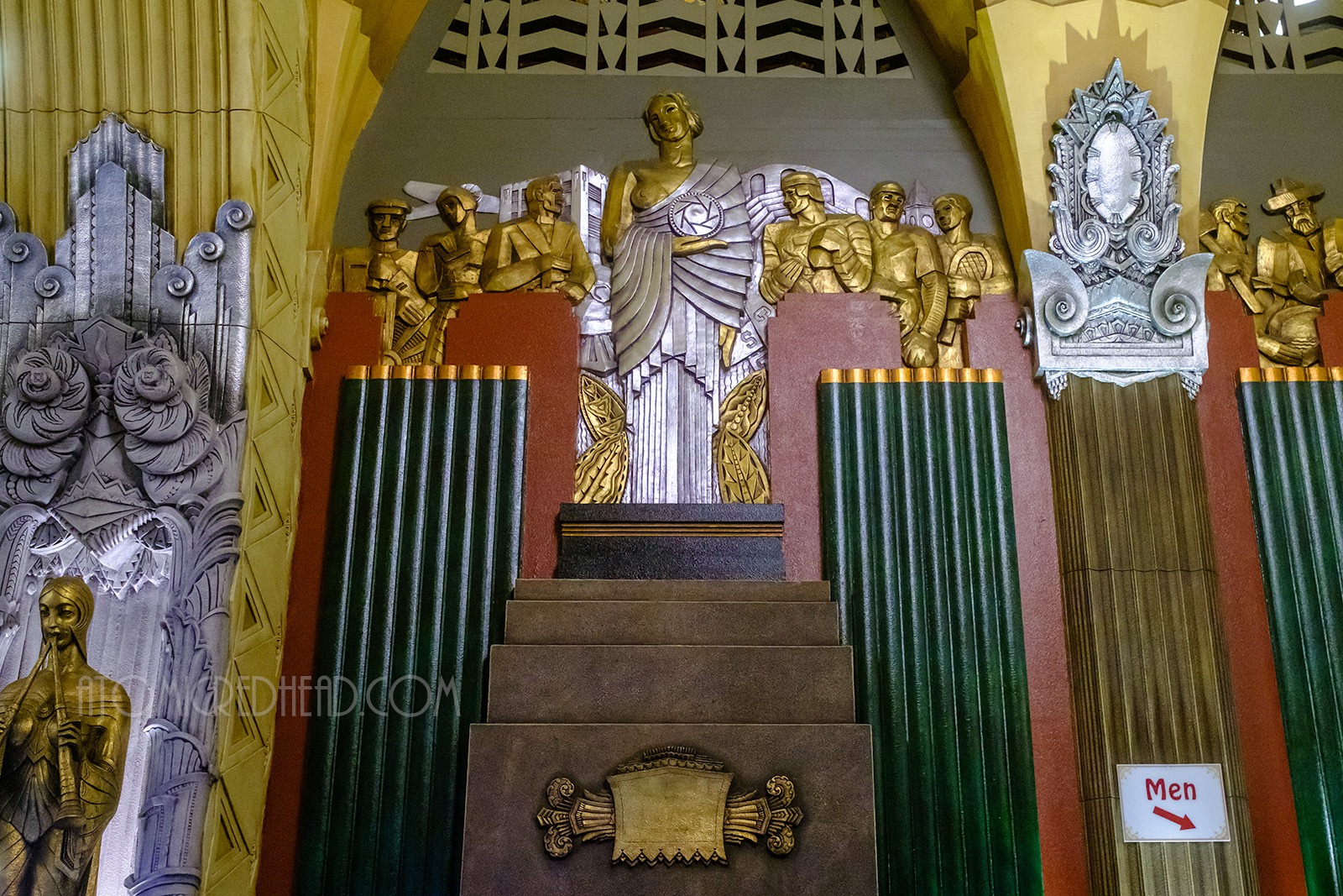 An Art Deco relief showing a silver woman holding a disc that has a shutter like image on it, behind her gold figures show athletes, and workers. Other Art Deco decorative elements surround her in shades of red, green and tan.