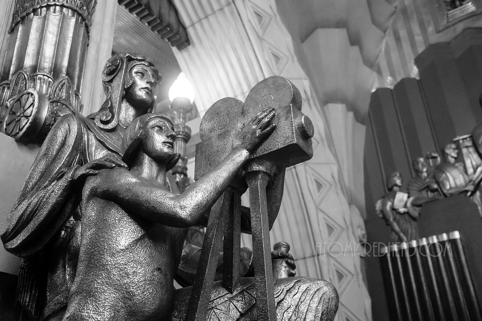 Black and white image of life size Art Deco statues, one has their hands on a film camera, the other stands behind with their hands on the shoulders of the cameraman.