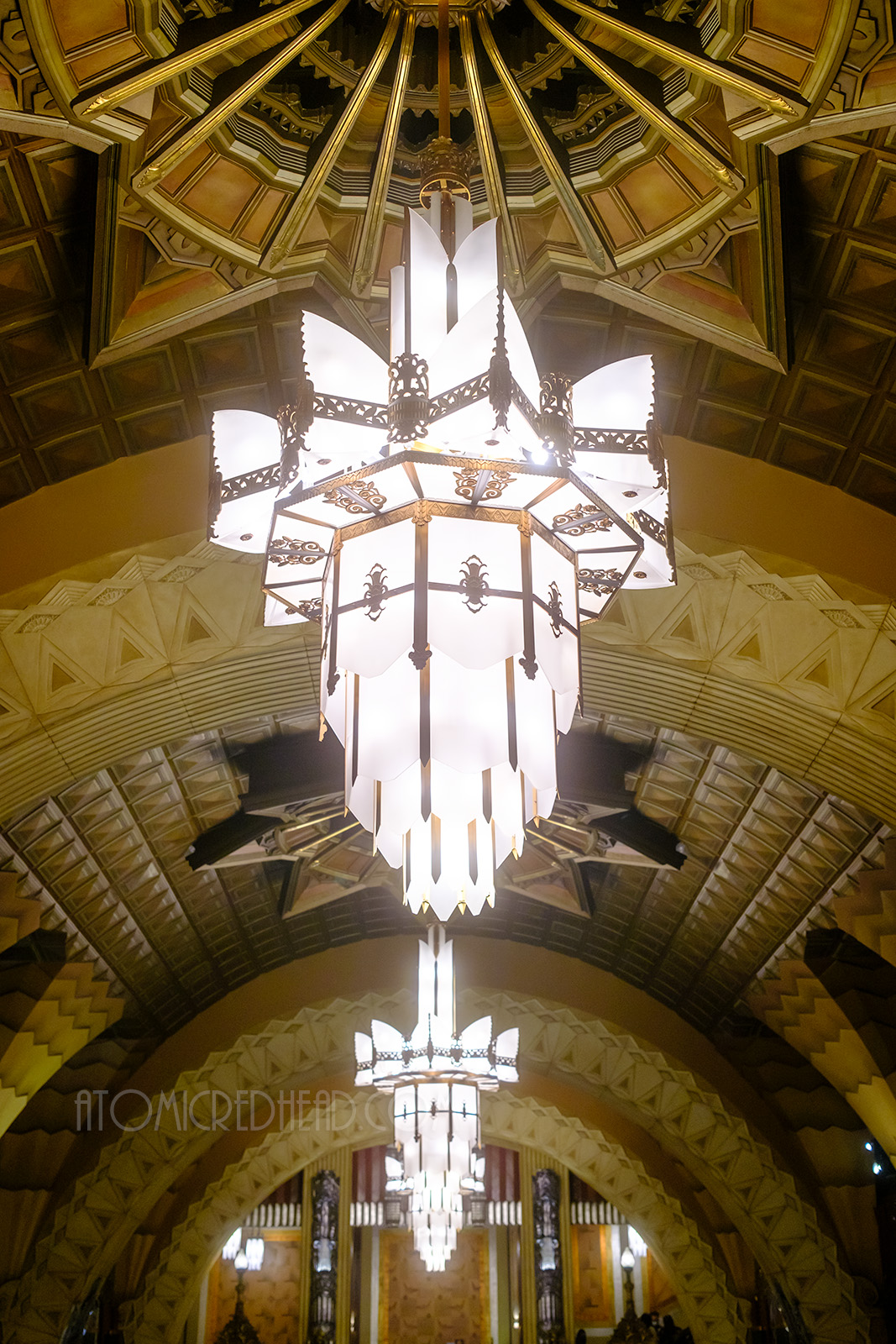 Three gold and white Art Deco chandeliers hang from an arched ceiling of the lobby. Various Art Deco shapes of triangles, diamonds and more make up a complex artistic ceiling.