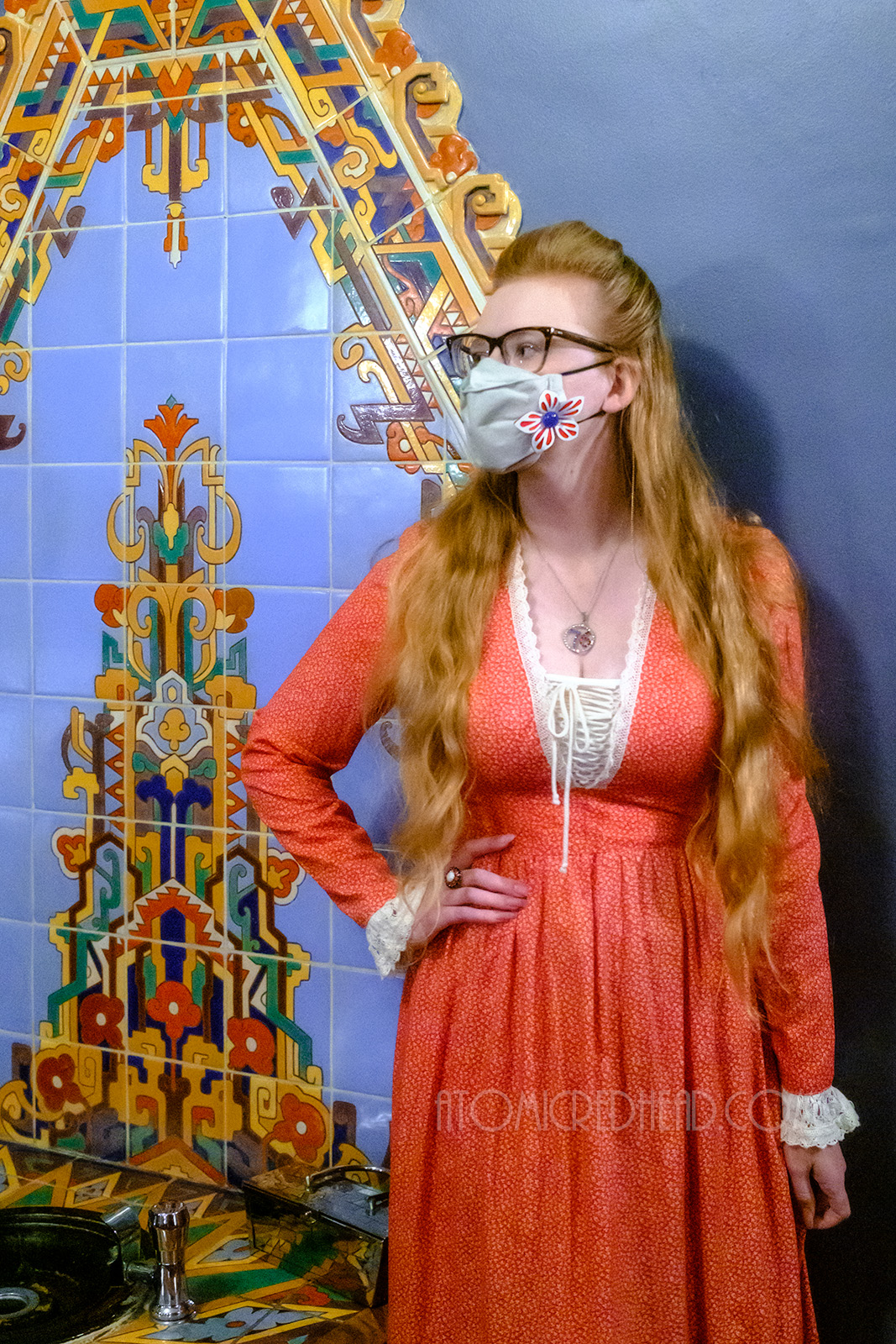Myself, wearing a red maxi length dress with a small print of tiny leaves, standing in front of an Art Deco tiled mural, with a design of blue, gold, green and red.