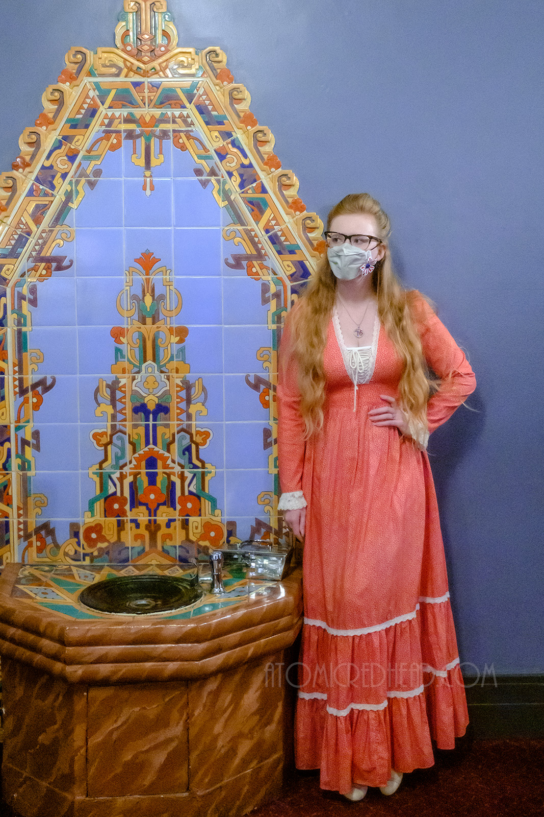 Myself, wearing a red maxi length dress with a small print of tiny leaves, standing in front of an Art Deco tiled mural, with a design of blue, gold, green and red.