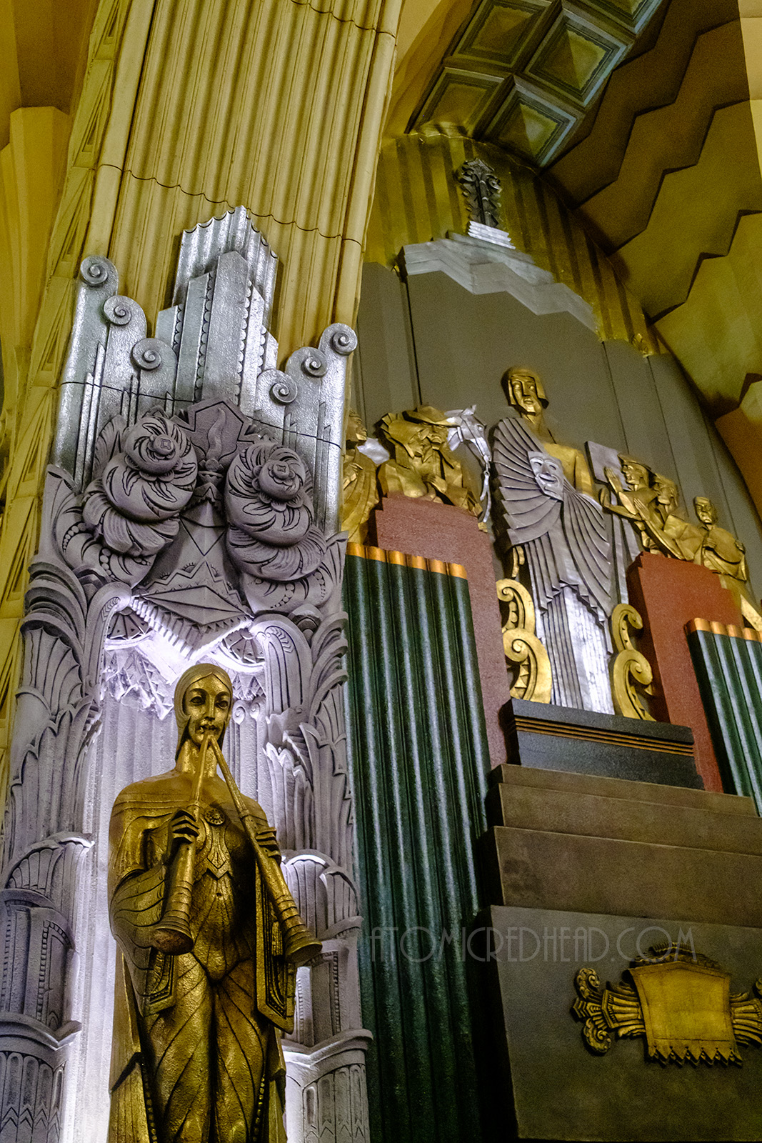 Looking up at a statue of a golden maiden with two horns in her hands. Behind her another silver maiden holds a mask, behind her gold figures appear to be filming a western film.