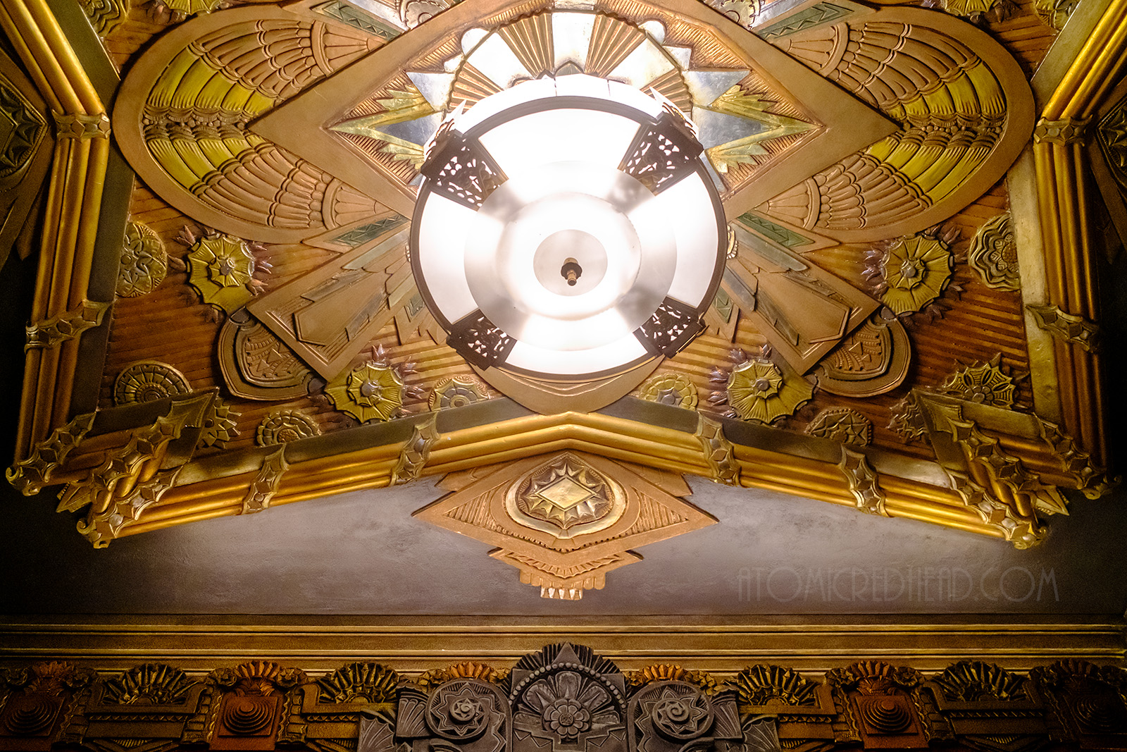 A massive round chandler hangs from a highly detailed and textured ceiling of Art Deco designs featuring swirls and sharp angles. 